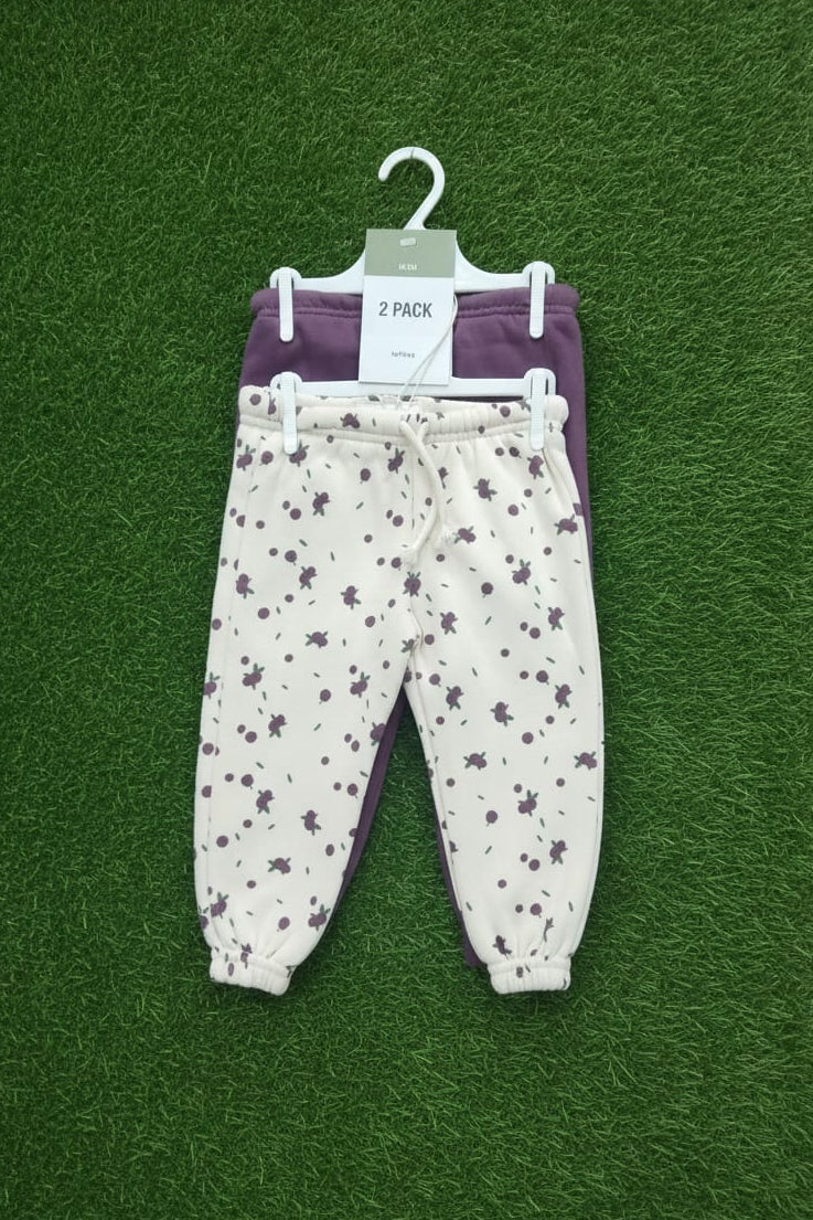 Two pairs of baby pants, one white with purple floral pattern and one purple, on a green background.